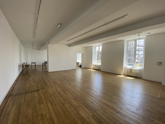 Grande salle vide avec plancher en bois, éclairage encastré, fenêtres et radiateur.