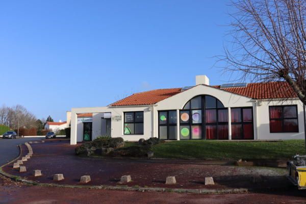 Bâtiment blanc avec toiture rouge, grandes fenêtres, parking et végétation.