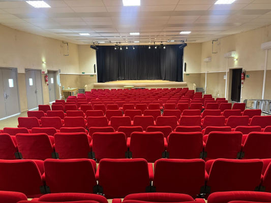 Salle avec rangées de sièges rouges, scène, rideau, éclairage et portes.