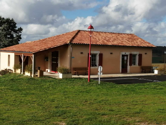 Maison basse avec toiture en tuiles rouges, portique, lampadaire et bancs près d'une aire de stationnement.