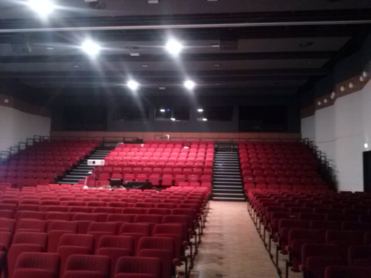 Salle de spectacle avec sièges rouges, scène et écran.