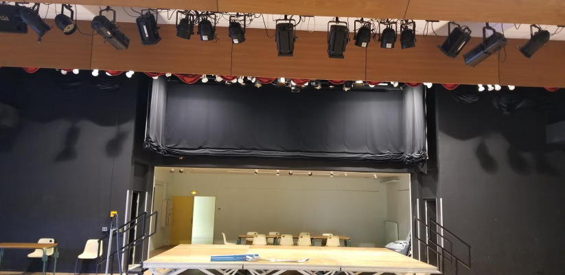 Scène avec projecteurs suspendus, fond noir, table et chaises.