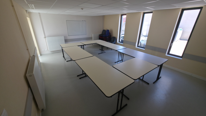 Salle avec tables, chaises, tableau blanc, radiateur et fenêtres.
