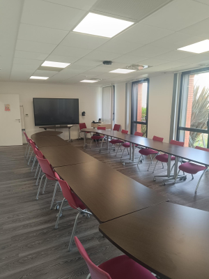 Salle de réunion avec tables, chaises, écran et fenêtres.