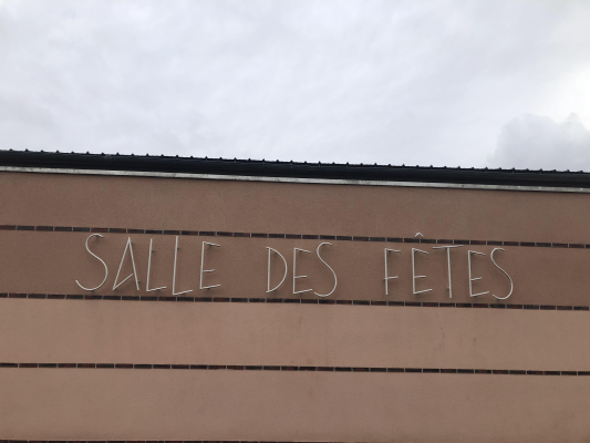 Image d'une enseigne sur un mur extérieur disant "Salle des fêtes" sous un ciel nuageux.