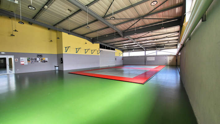 Salle de sport avec sol vert, tapis rouge, murs jaunes et gris. Haut plafond métallique.