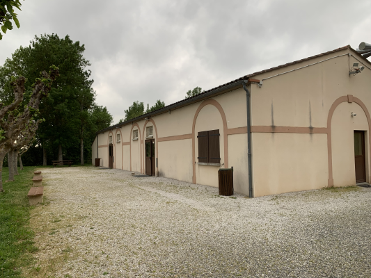 Bâtiment rectangulaire avec murs beiges, portes et fenêtres sombres, gravier au sol, poubelle extérieure et arbre à gauche.