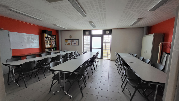 Salle avec tables, chaises, tableau blanc, étagères, porte et fenêtres.