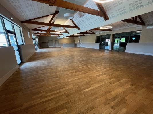 Grande salle avec parquet, poutres apparentes, luminaires suspendus, portes vitrées et fenêtres.