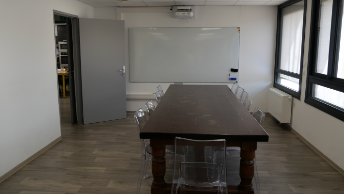 Salle avec table, chaises, tableau blanc, vidéo projecteur et radiateur.