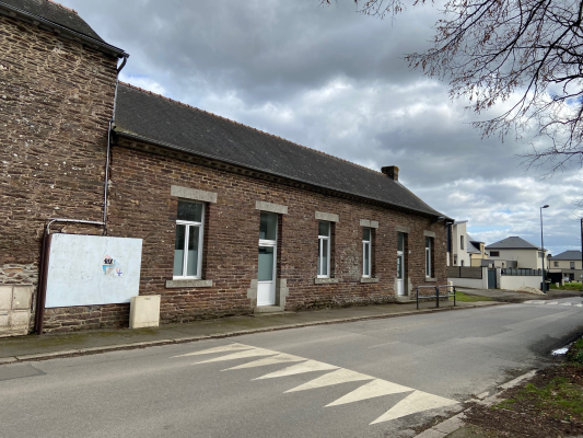 Un bâtiment en brique à un étage avec des fenêtres blanches, un panneau, une route avec un passage piéton, des arbres sans feuilles.