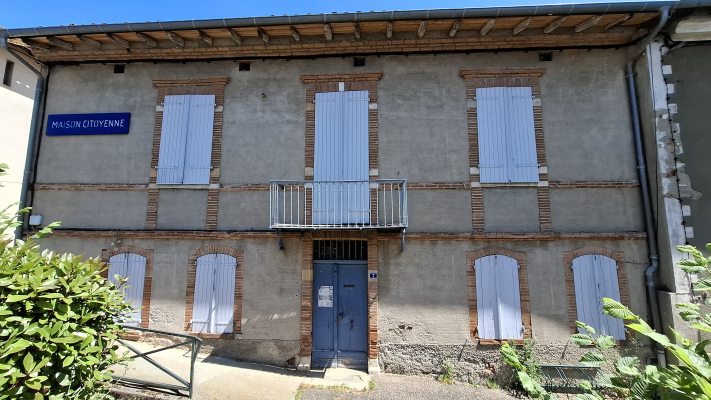 Bâtiment avec façade en briques, volets gris, balcon métallique, porte bleue et panneau "Maison Citoyenne".