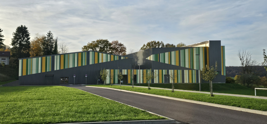 Bâtiment moderne avec façades striées en gris, jaune et vert. Allée, pelouse et arbres.