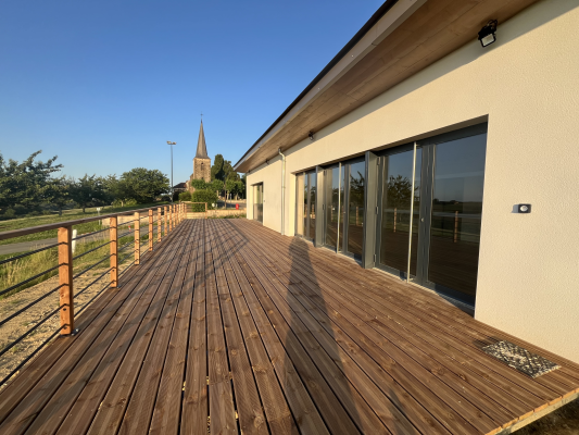 Terrasse en bois menant à un bâtiment avec fenêtres vitrées. Eglise lointaine et ciel dégagé.