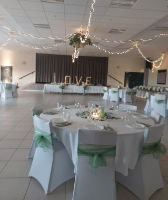 Salle de réception avec tables, chaises, nappes, vaisselle, guirlandes lumineuses et décorations florales.