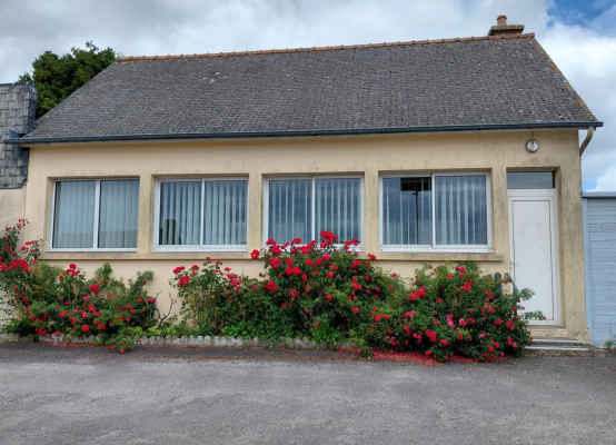 Maison beige avec toit gris, trois fenêtres, porte blanche, et buissons de fleurs rouges.