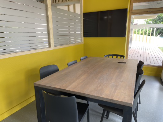 Table de réunion avec six chaises, mur jaune, fenêtres avec stores et porte ouverte.