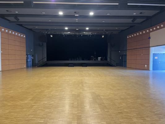 Salle vaste avec plancher en bois, scène, éclairages, murs en bois et portes bleues.