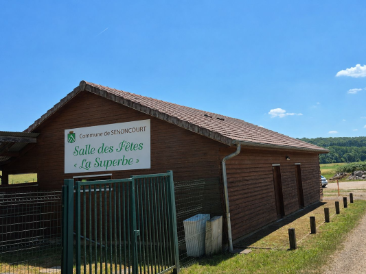 Bâtiment en bois avec une pancarte indiquant "Salle des Fêtes La Superbe" et un portail vert.