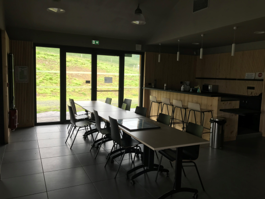 Salle avec tables, chaises, cuisine et grande porte vitrée.