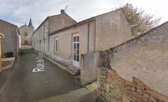 Rue étroite avec bâtiments anciens en pierre, église au fond, poteaux électriques, arbres dénudés.