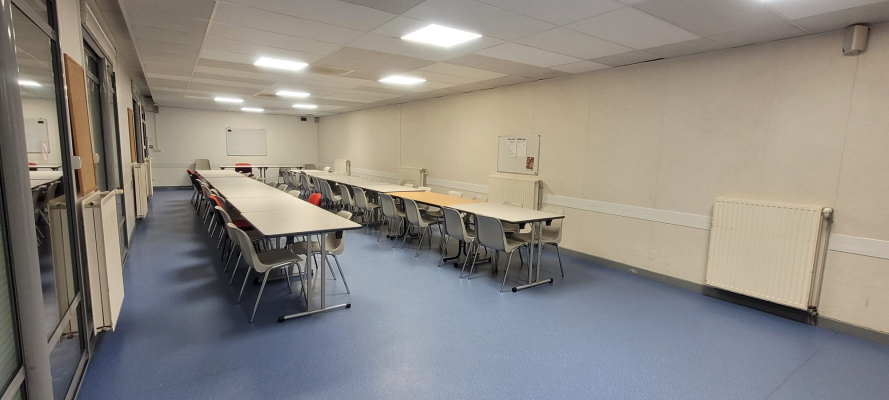 Salle de classe avec tables, chaises, tableau blanc et radiateurs.