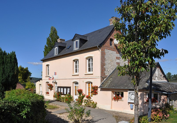 Maison à deux étages avec toit noir, murs beiges et pierres apparentes. Fenêtres avec volets blancs. Jardinières avec fleurs rouges. Arbre à droite. Ciel bleu.