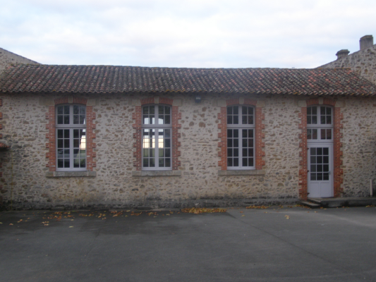 Bâtiment en pierre avec toiture en tuiles, quatre fenêtres et une porte blanche. Cour asphalte.