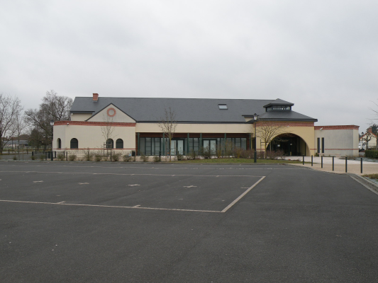 Bâtiment avec façade beige, toiture grise, fenêtres et porte d'entrée. Parking vide devant.