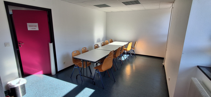 Salle avec tables, chaises, porte rouge, radiateur et poubelle.
